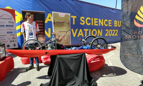 Zdjęcie przed budynkiem Centrum Nauki i Techniki EC1, mężczyzna prezentuje pojazd kołowy o napędzie elektrycznym, stoisko – LODZ SOLAR TEAM, za mężczyzną widnieje baner reklamowy targów.